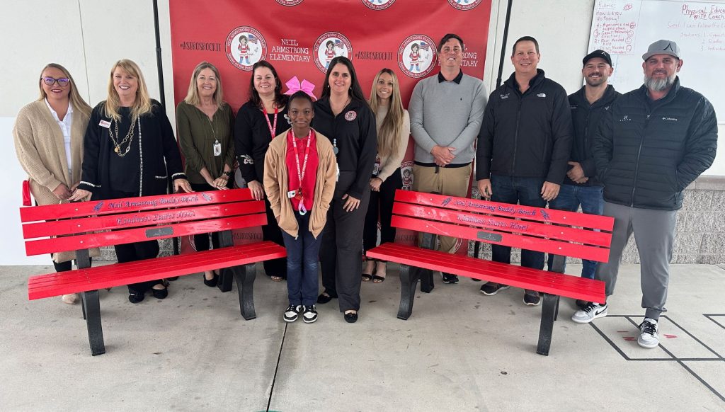 Alair Homes team members stand beside two custom-designed 'Buddy Benches' at Neil Armstrong Elementary. The benches, presented to the school as part of Alair's community initiative, create a welcoming space for students to connect and build friendships during recess.